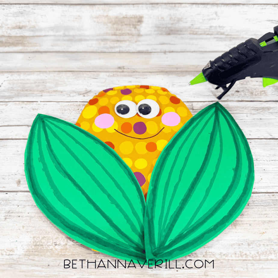 Dotted paper plate corn face positioned between two large green paper leaves with a hot glue gun nearby.