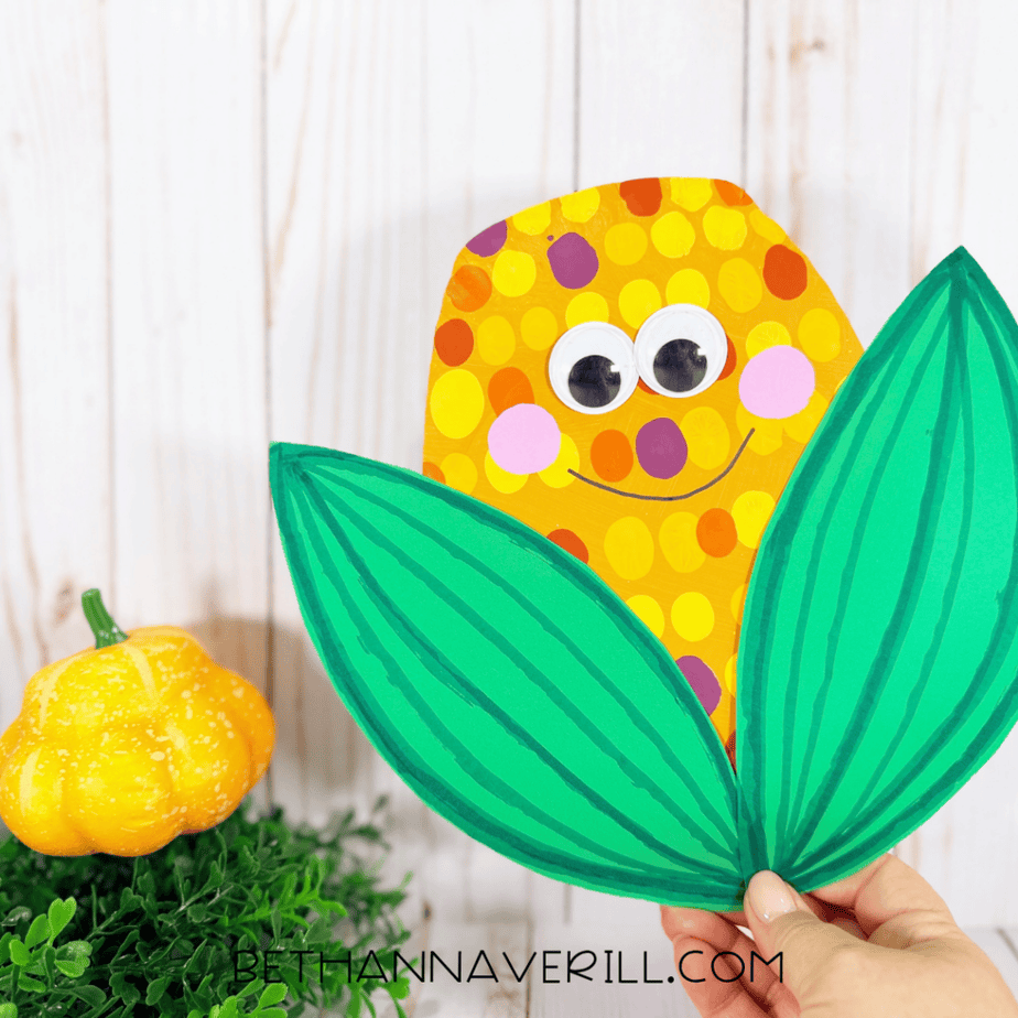 Finished harvest corn paper plate craft with colorful painted dots, googly eyes, pink cheeks, smiling face, and two large green leaves, held upright beside a small yellow pumpkin.
