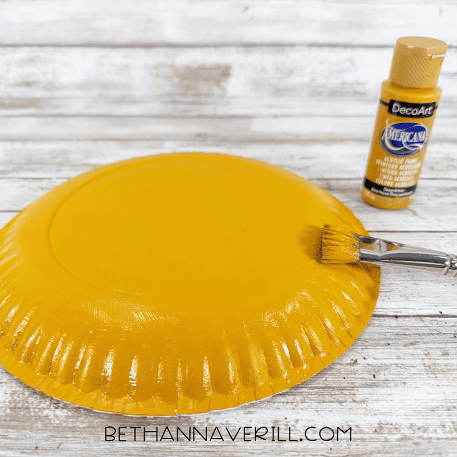 Paper plate painted golden yellow beside a yellow acrylic paint bottle and paintbrush on a white wood surface.
