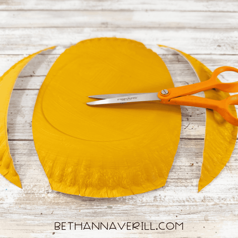 Golden yellow painted paper plate trimmed into an oval shape with curved scraps on both sides and orange scissors resting on top.