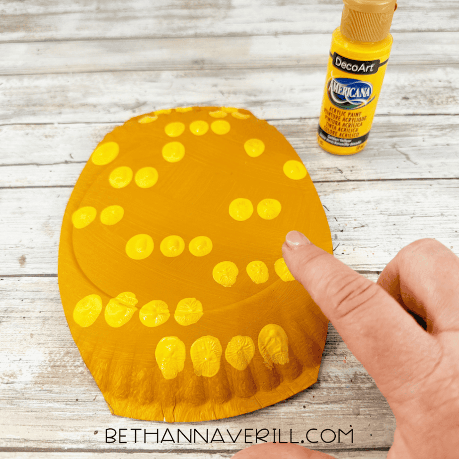 Finger pressing bright yellow paint dots onto a golden yellow paper plate with a yellow acrylic paint bottle nearby.