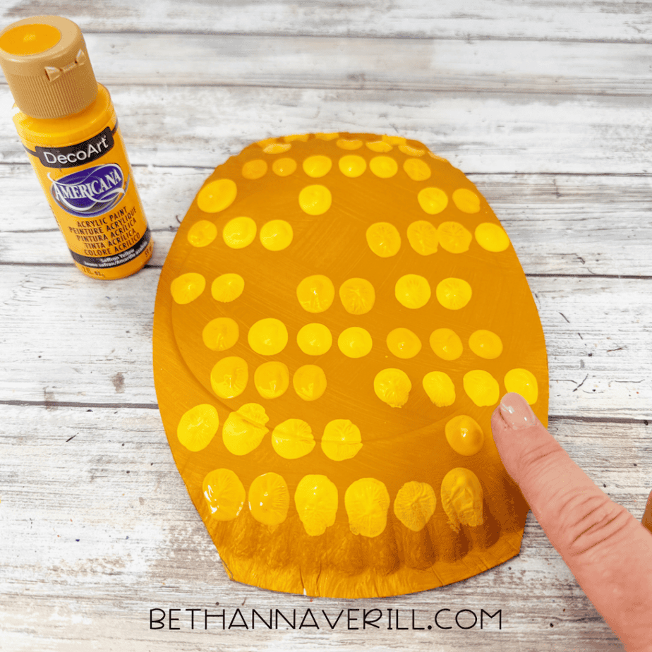 Golden yellow paper plate covered with rows of bright yellow paint dots as a finger adds another dot.