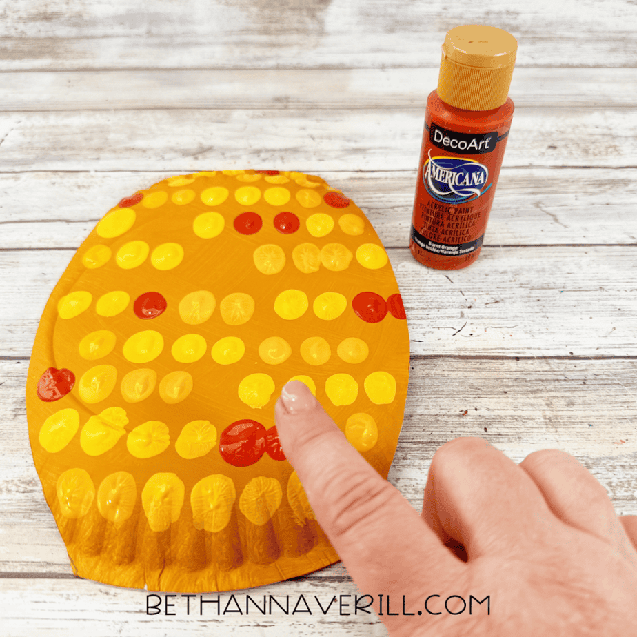 Golden yellow paper plate with yellow and orange paint dots as a finger adds orange paint, with an orange acrylic paint bottle nearby.
