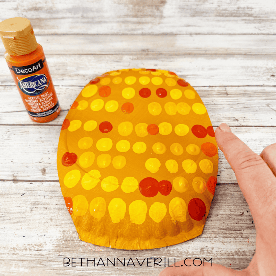 Golden yellow paper plate covered with yellow and orange paint dots while a finger adds another orange dot.