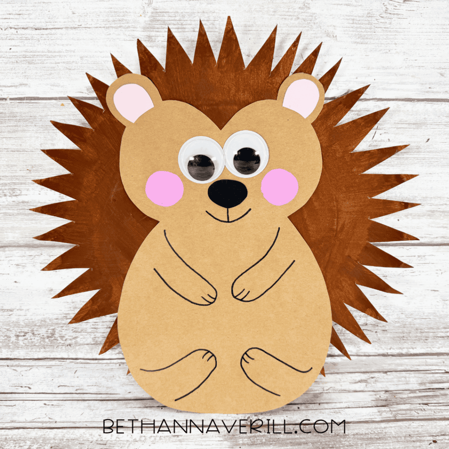 Finished paper plate hedgehog with a tan face and body, brown pointed paper plate spines, googly eyes, pink cheeks and inner ears, black nose, and black outlined arms and feet.