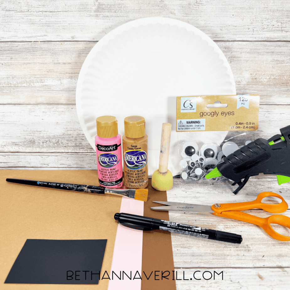 White paper plate with brown and pink acrylic paint, paintbrush, foam dauber, googly eyes, hot glue gun, orange scissors, black marker, and sheets of tan, brown, pink, and black paper.