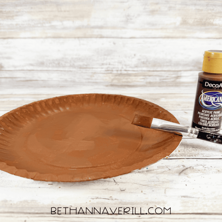Paintbrush applying brown acrylic paint to a paper plate, with a brown paint bottle beside it.