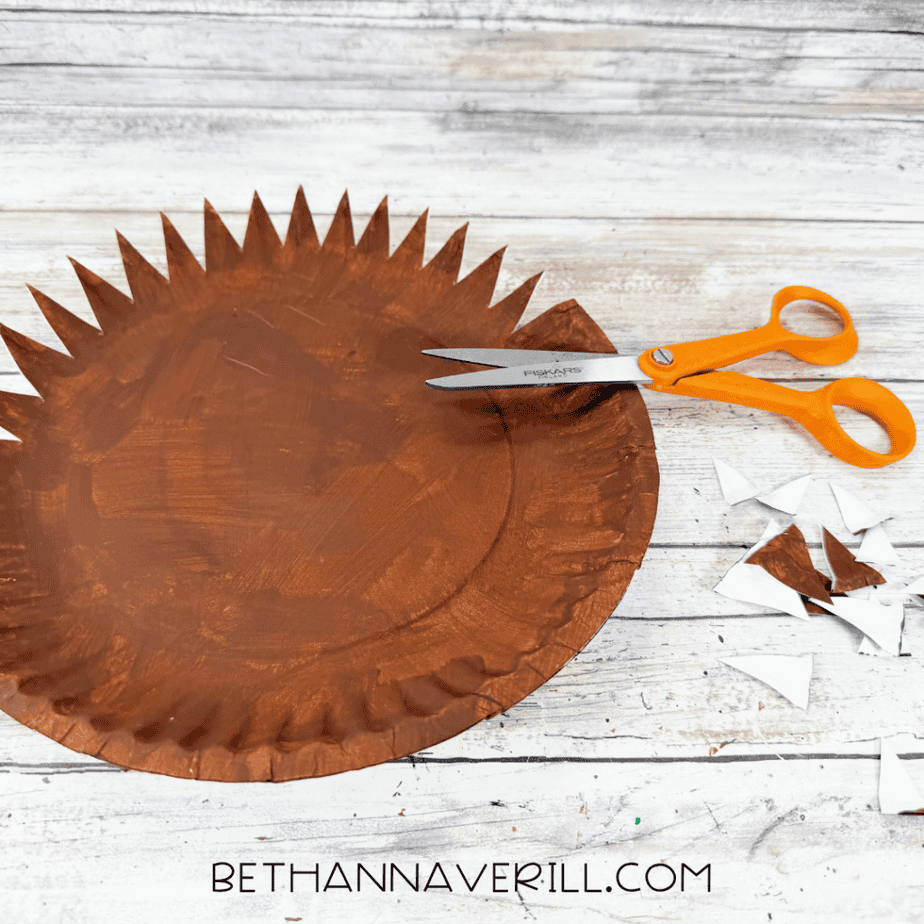Brown painted paper plate with pointed sections cut around the edge, with orange scissors and paper scraps nearby.