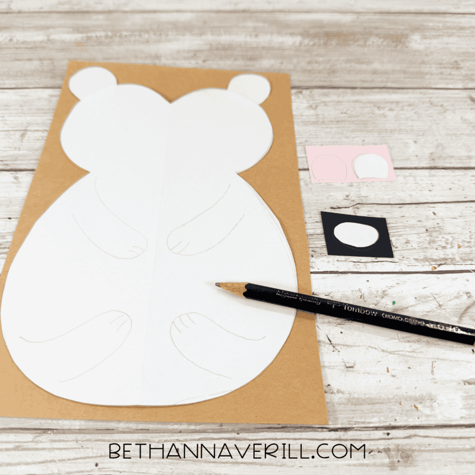 White paper template shaped like a rounded animal body positioned on tan paper, with smaller pink and black paper pieces and a black pencil nearby.
