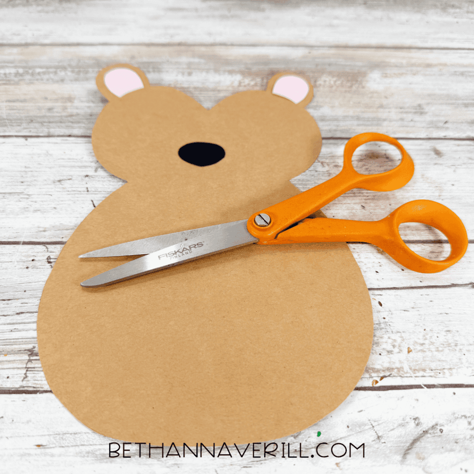 Cut tan paper animal shape with pink inner ears and a black oval nose, with orange scissors resting across the body.
