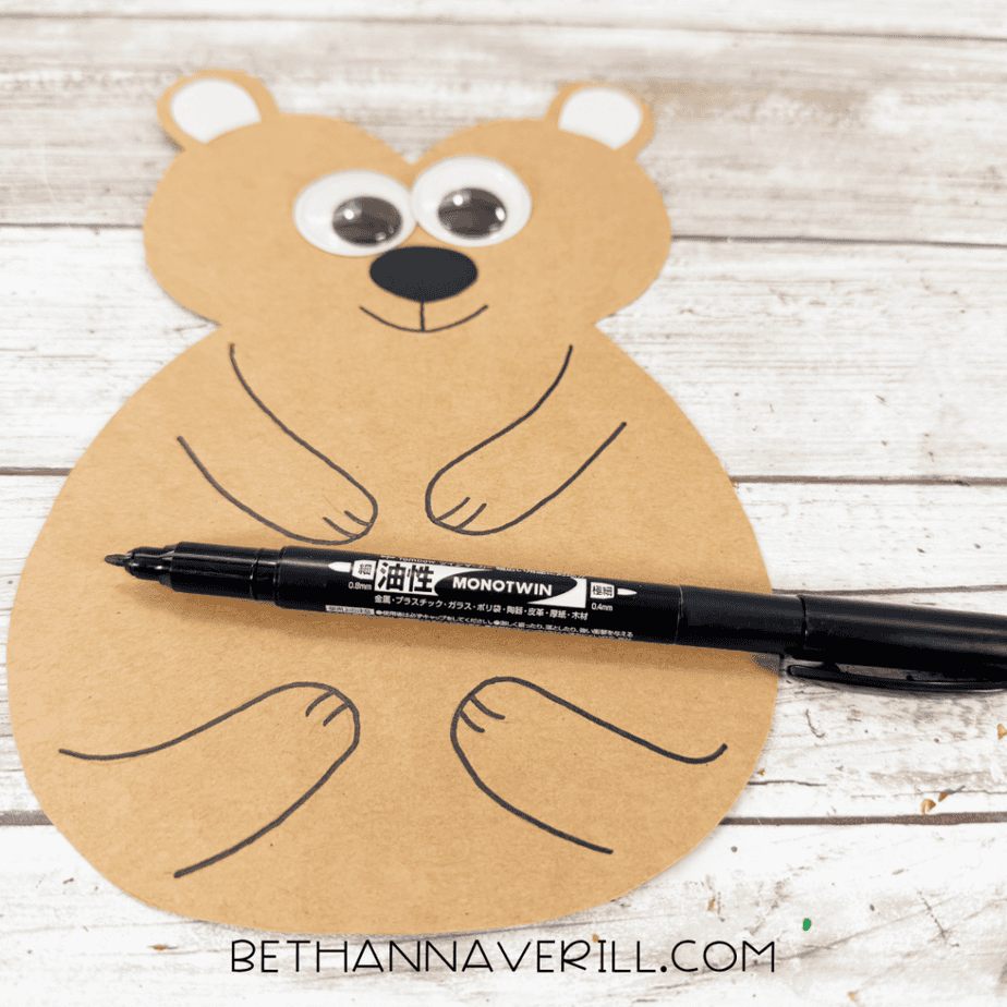 Tan paper animal shape with googly eyes, black nose, and outlined arms, legs, and mouth, with a black marker resting across the body.