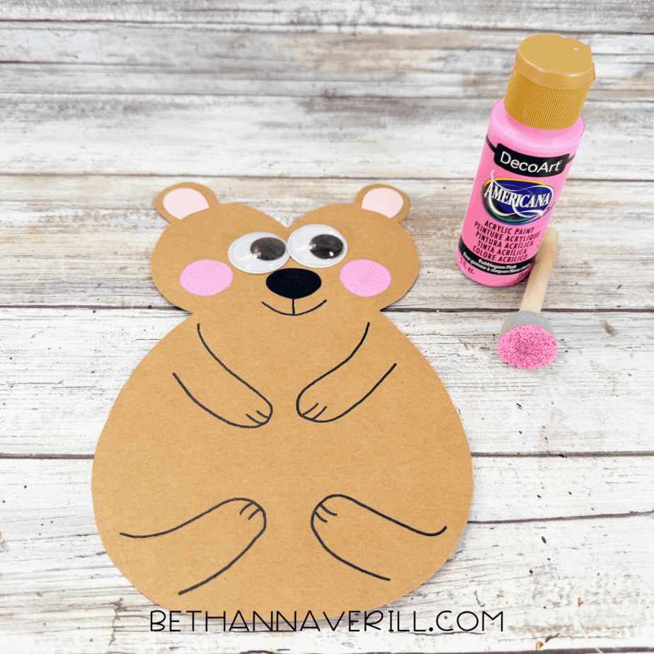 Tan paper animal shape with pink painted cheeks, googly eyes, black nose, and black outlined body details, beside pink paint and a foam dauber.