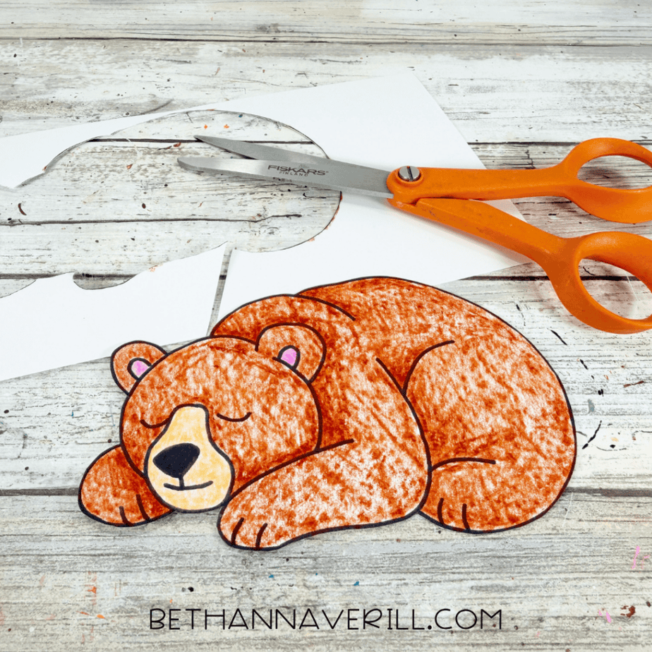 Cut-out colored sleeping bear lying on a white wood surface with orange-handled scissors and paper scraps nearby.