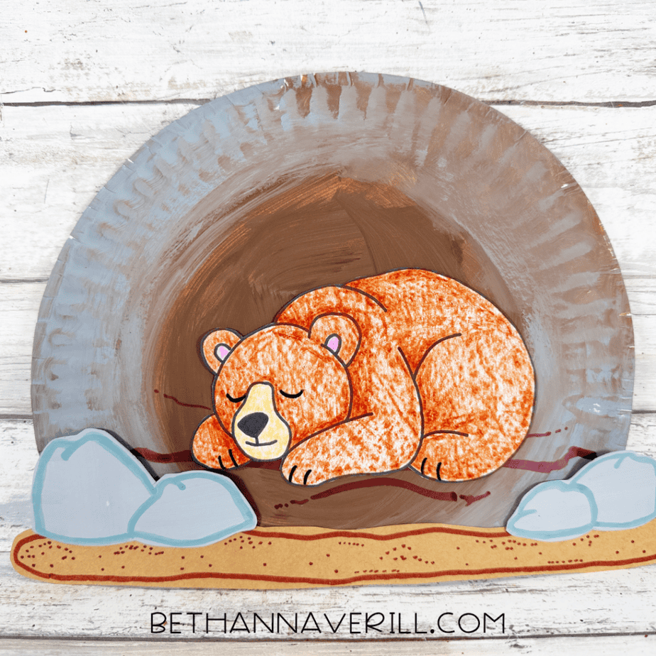 Completed hibernating bear paper plate craft with a sleeping brown bear inside a painted cave, gray rocks on both sides, and a brown ground strip across the bottom.