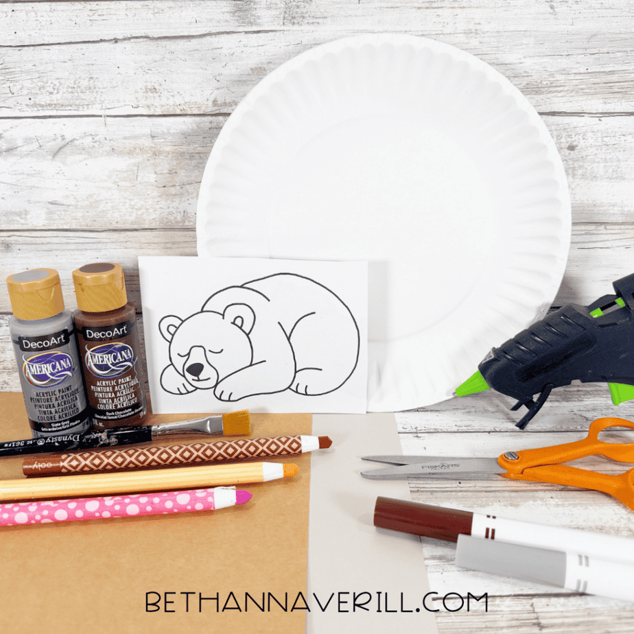 White paper plate with gray and brown acrylic paint, paintbrush, colored pencils, markers, scissors, hot glue gun, cardstock, and a printed sleeping bear drawing arranged on a white wood surface.