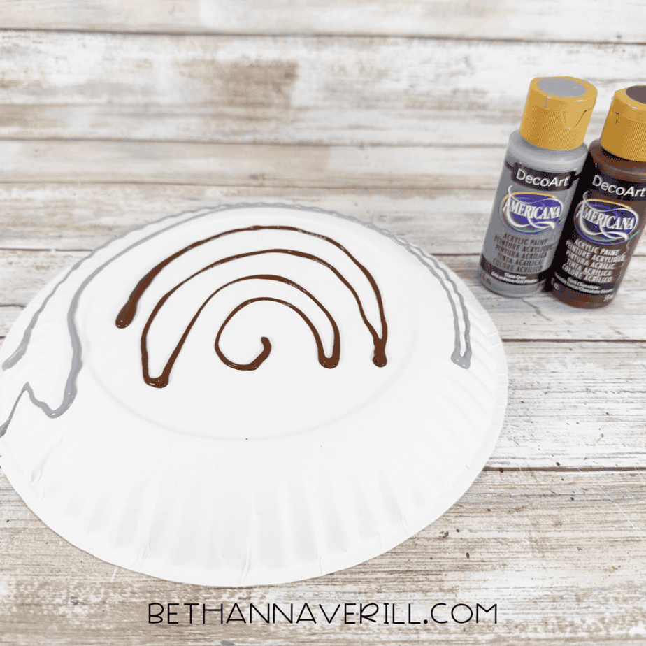 White paper plate with curved lines of brown and gray paint across the back, with two acrylic paint bottles beside it.