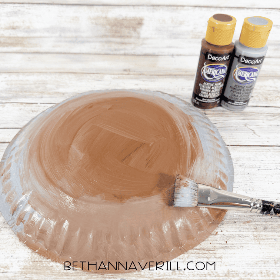 Paper plate being painted with blended brown and gray acrylic paint using a flat paintbrush.
