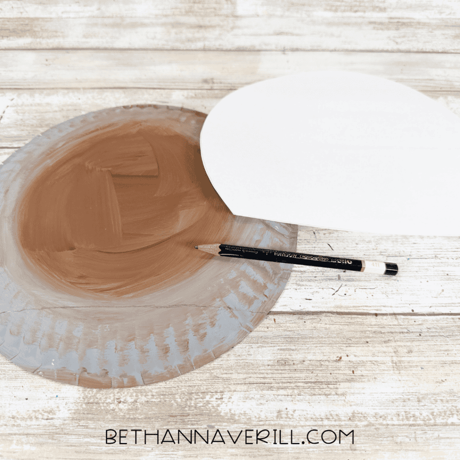 Painted paper plate beside a white paper template while a pencil traces a curved shape onto the plate.
