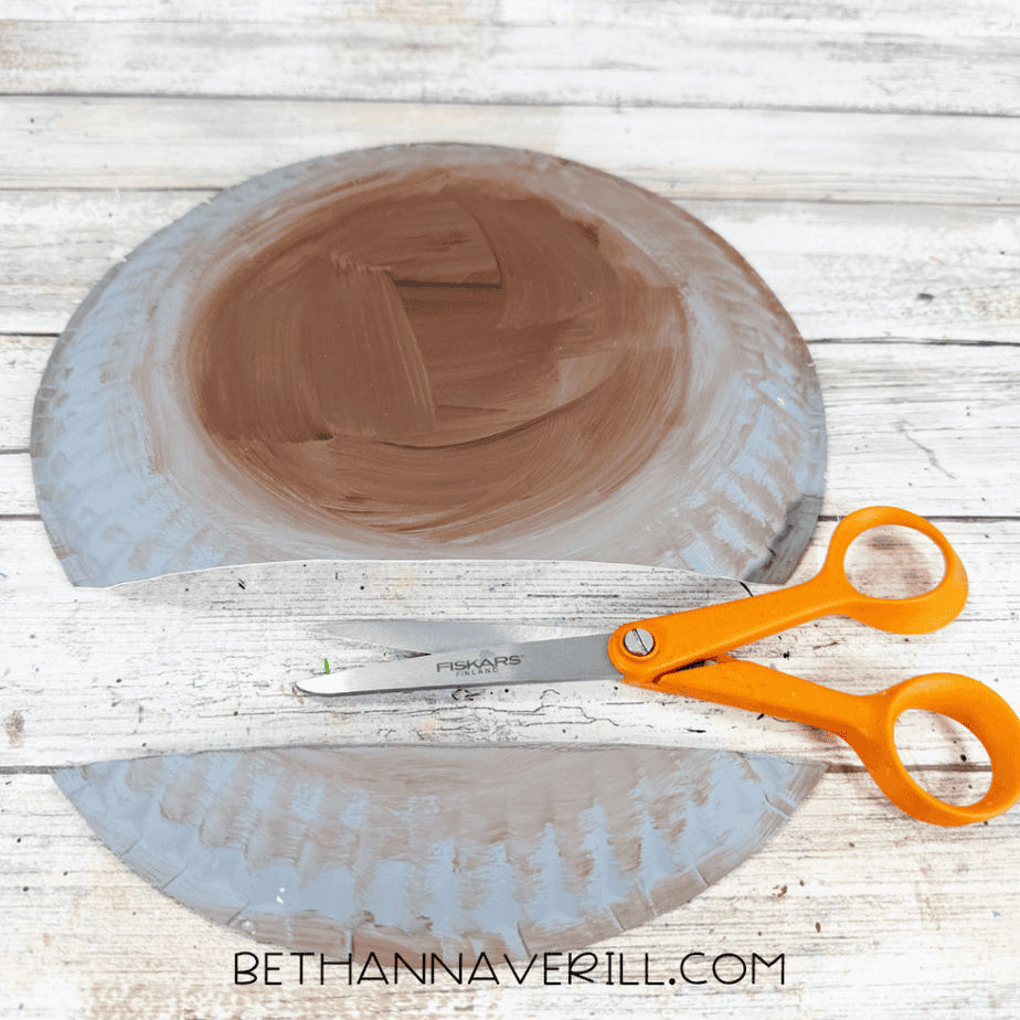 Painted paper plate cut into a large rounded section and a smaller curved strip, with orange-handled scissors nearby.