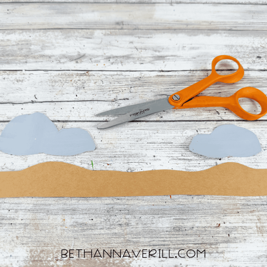 Two sets of gray paper rock shapes and a long brown wavy paper strip arranged beside orange-handled scissors.