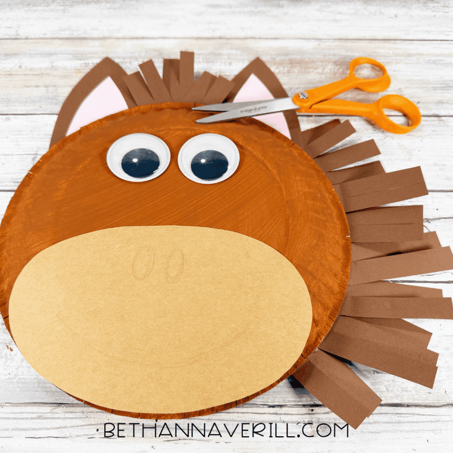 Paper plate horse face with many brown paper strips around one side, googly eyes, pink-centered ears, and a tan muzzle, with orange scissors above.