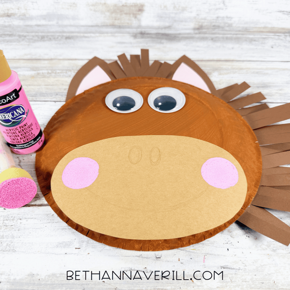 Paper plate horse face with pink circles painted on the tan muzzle, beside a pink acrylic paint bottle and foam dauber.