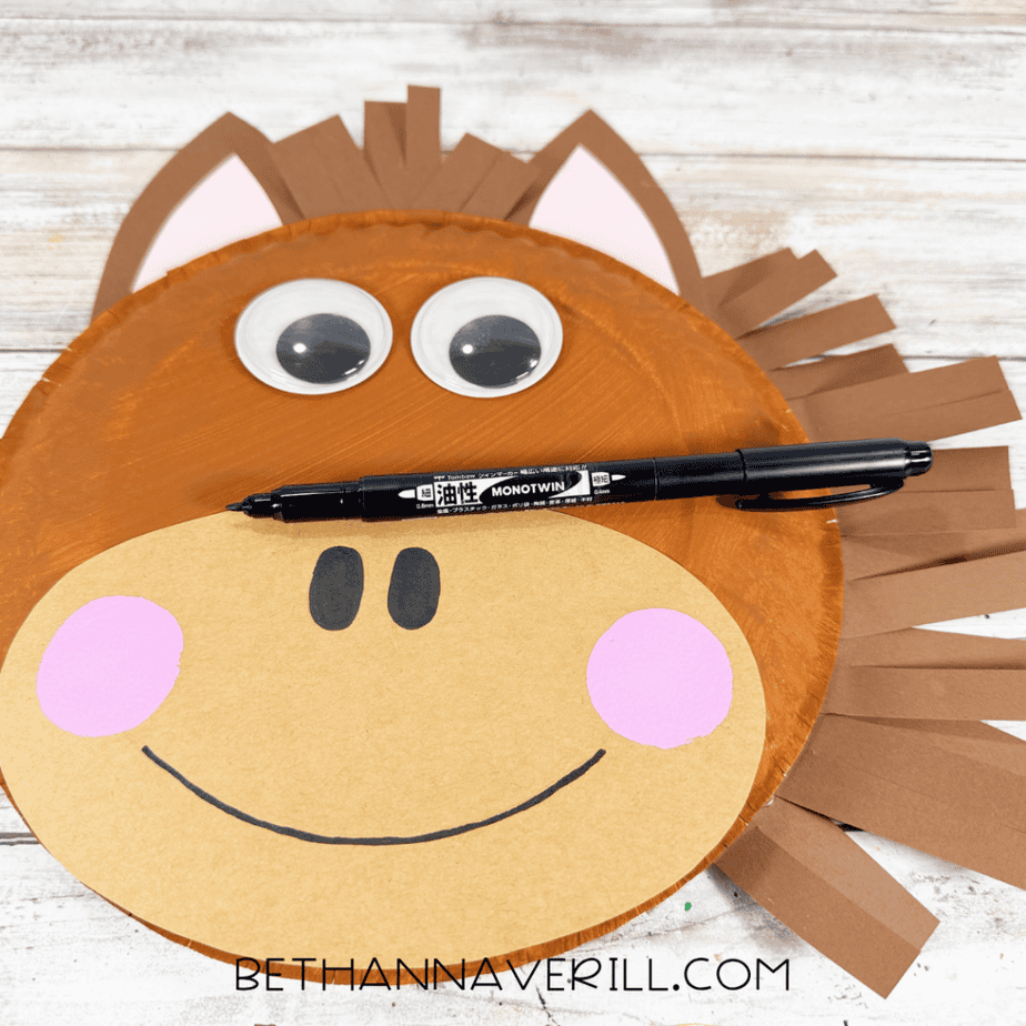Paper plate horse face with googly eyes, brown paper mane, pink cheeks, black nostrils, and a curved black smile, with a black marker across the face.