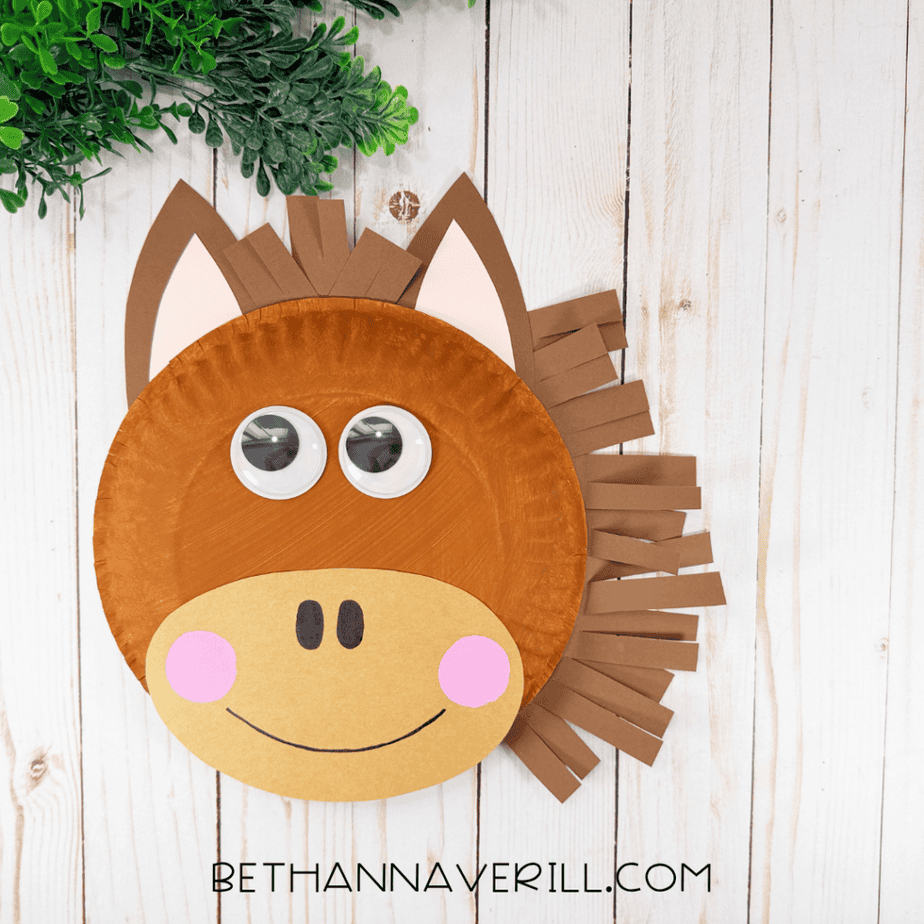 Finished paper plate horse face with brown and tan coloring, pink-centered ears, googly eyes, pink cheeks, black nostrils, a smiling mouth, and brown paper strips forming the mane.