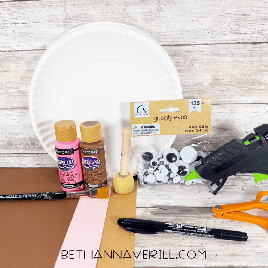 White paper plate with pink and brown acrylic paint, foam dauber, paintbrush, googly eyes, hot glue gun, orange scissors, black marker, and sheets of brown, tan, and pink paper.