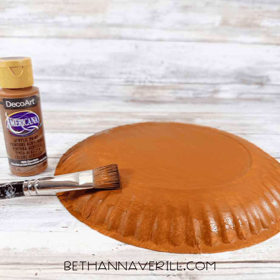 Paintbrush applying brown acrylic paint to the back of a paper plate, with a brown paint bottle beside it.