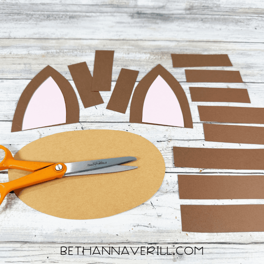 Cut paper pieces arranged on a white wood surface, including two brown ears with pink centers, a large tan oval, and several brown rectangular strips, with orange scissors nearby.