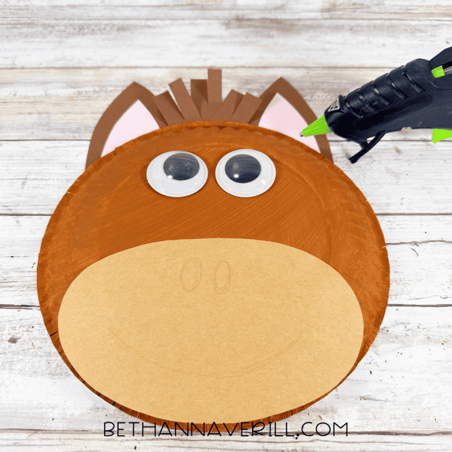 Partially assembled paper plate horse face with brown ears, brown paper strips at the top, large googly eyes, and a tan oval muzzle.