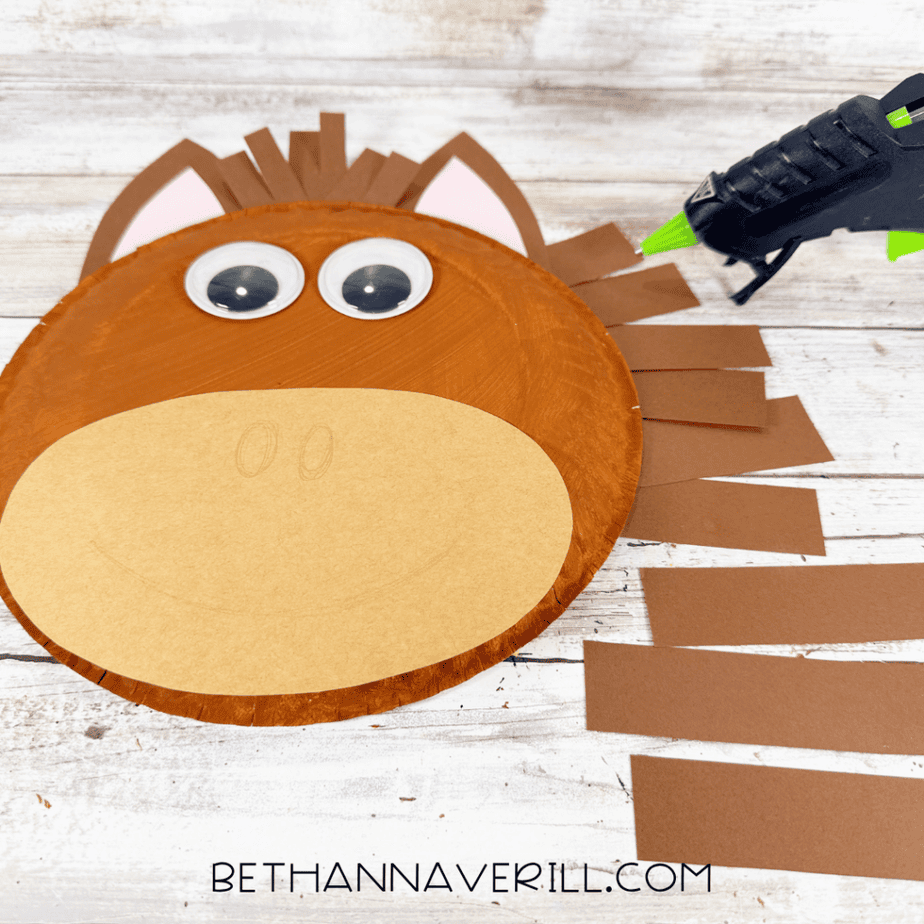Paper plate horse face with brown paper strips extending along one side, googly eyes, brown ears with pink centers, and a tan oval muzzle, with a hot glue gun nearby.