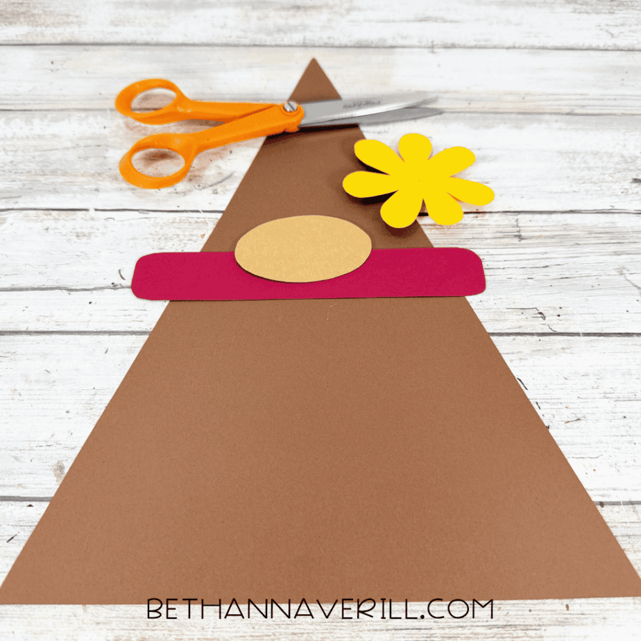 Brown triangular paper hat with red band, tan oval piece, yellow paper flower, and orange scissors arranged on a white wood surface.