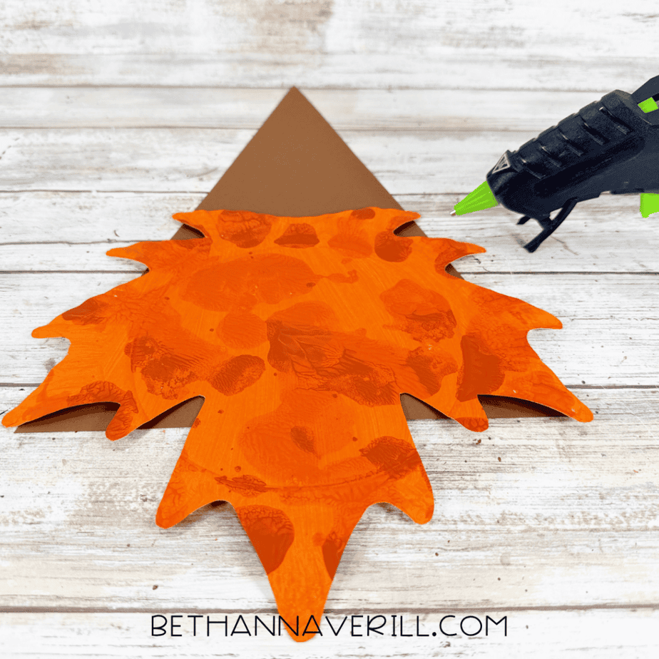 Orange leaf-shaped paper plate attached beneath a tall brown triangular hat with a red band, with a hot glue gun nearby.