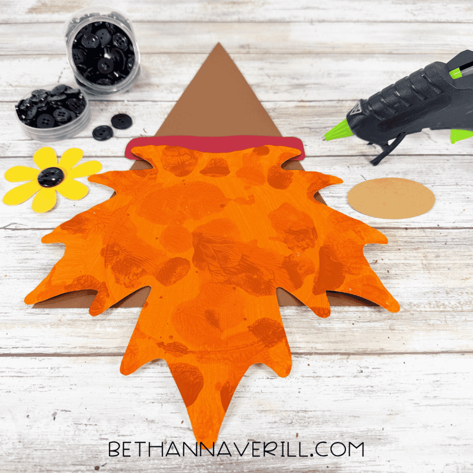Leaf gnome craft with orange textured paper plate beard, brown pointed hat, red hat band, tan oval nose, yellow flower, black buttons, and hot glue gun.