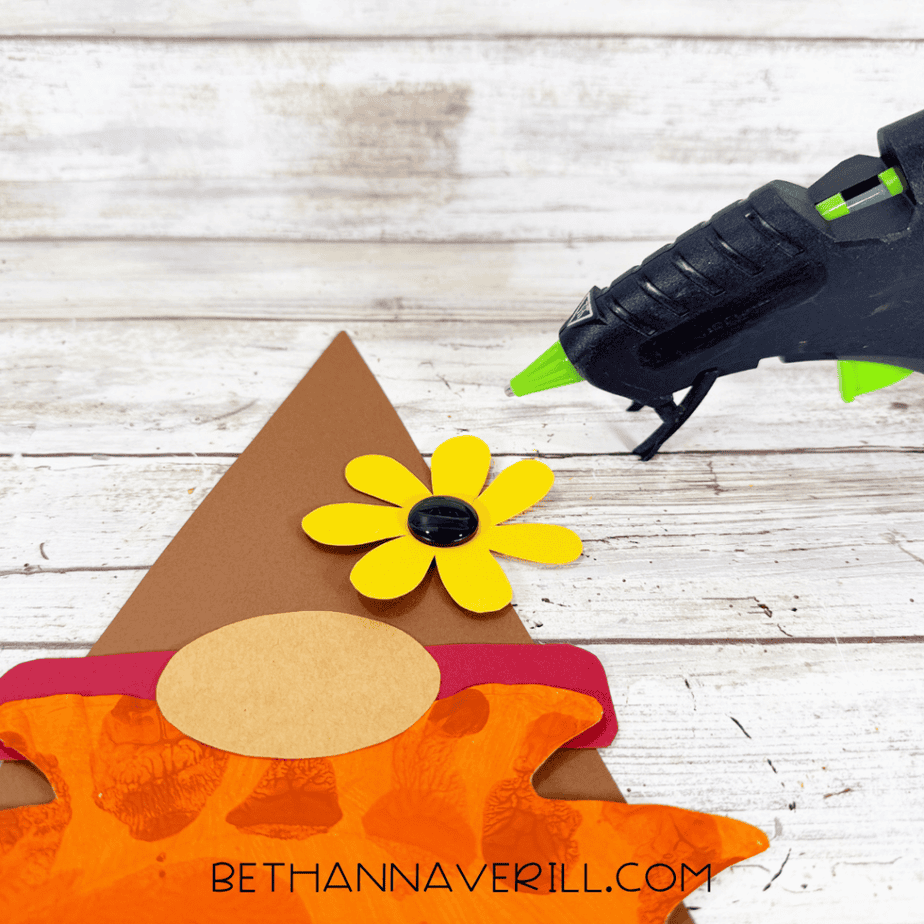 Close-up of a yellow paper flower with a black button center attached to the brown pointed hat of the leaf gnome craft.