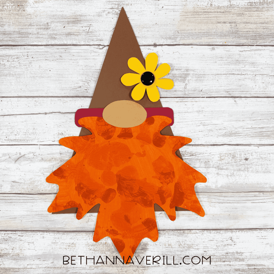 Finished leaf gnome craft with tall brown pointed hat, yellow flower with black button center, tan oval nose, red hat band, and orange textured paper plate beard.