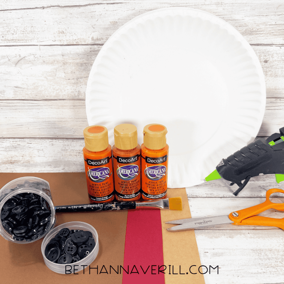 White paper plate with orange acrylic paint, brown and red paper, black buttons, paintbrush, hot glue gun, scissors, and craft supplies on a white wood surface.