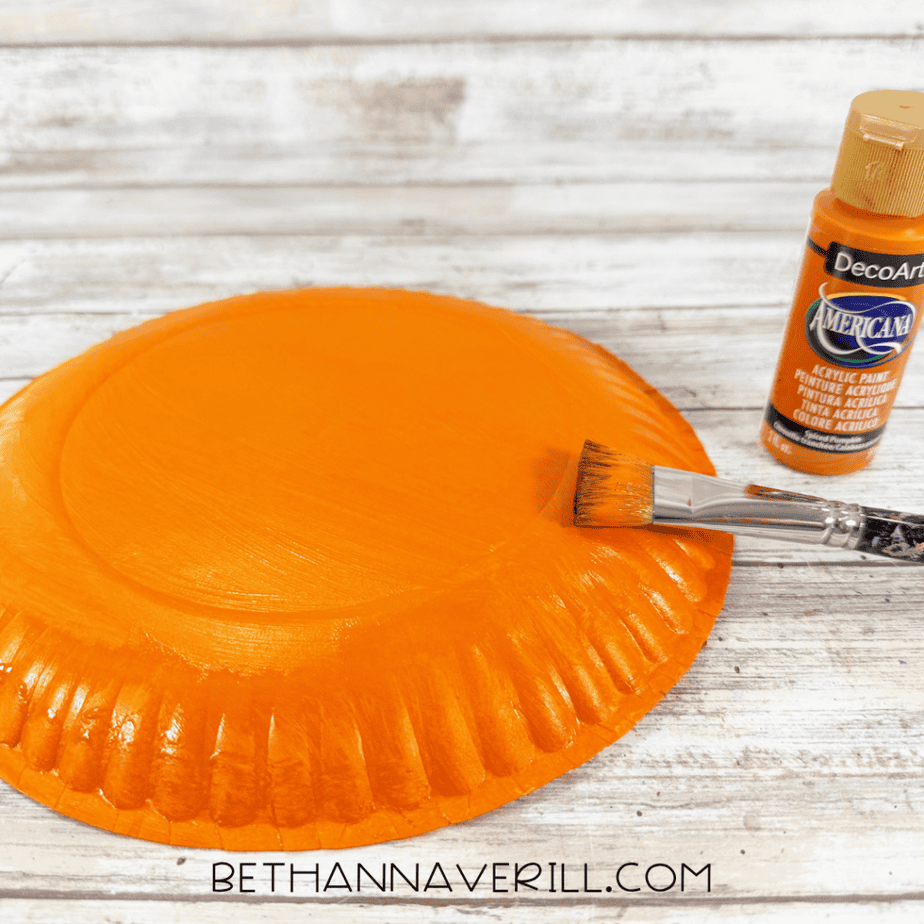 Paper plate painted bright orange with a paintbrush resting across the plate and orange acrylic paint beside it.