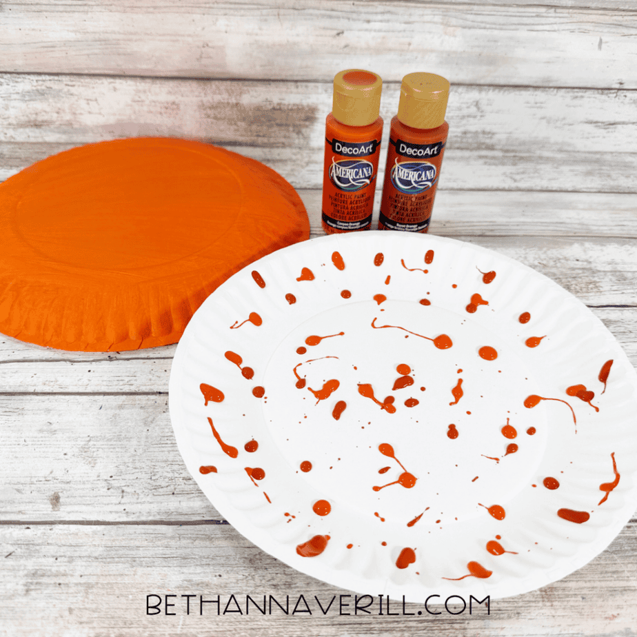 White paper plate covered with scattered orange paint marks beside an orange painted paper plate and two bottles of orange acrylic paint.