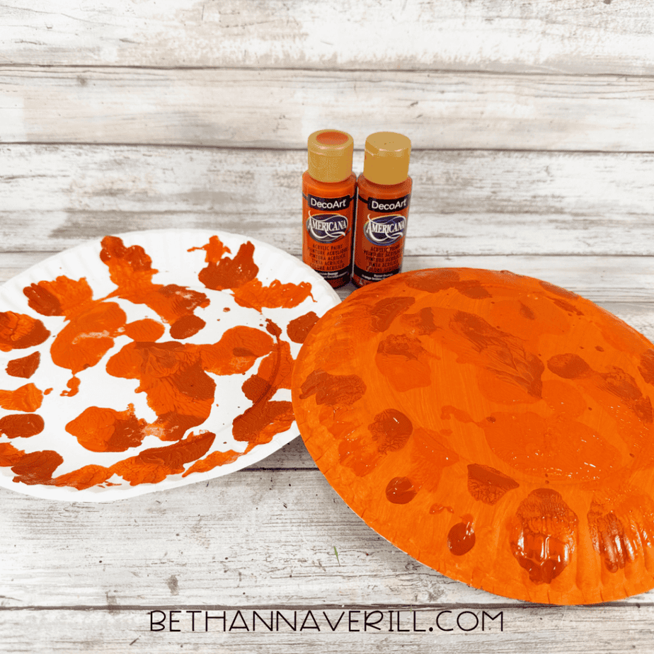 Orange painted paper plate with darker orange textured paint marks beside a white plate and two bottles of orange paint.