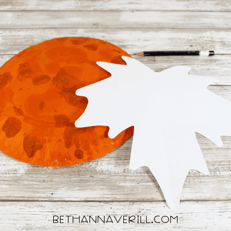 Orange painted paper plate beside a large white leaf-shaped paper template and a black pencil.