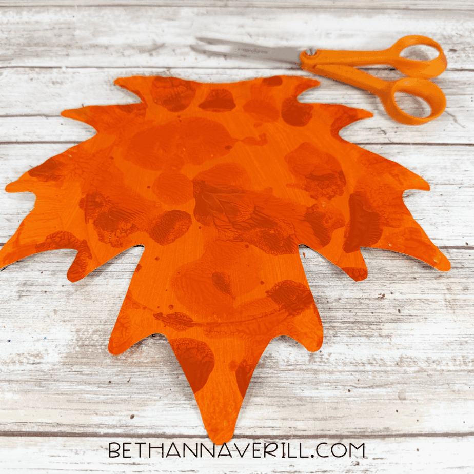 Orange paper plate cut into a pointed leaf shape with darker orange painted texture and orange scissors above it.