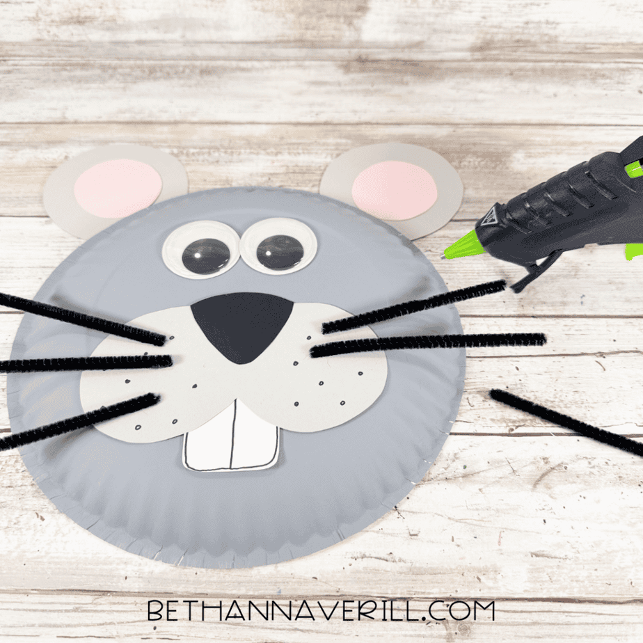 Gray paper plate mouse face with black pipe cleaner whiskers being positioned along both sides of the muzzle, with a hot glue gun nearby.