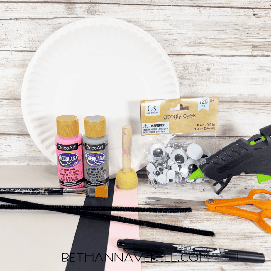 White paper plate with pink and gray acrylic paint, foam dauber, googly eyes, hot glue gun, orange scissors, black marker, black pipe cleaners, and sheets of gray, black, pink, and white paper.