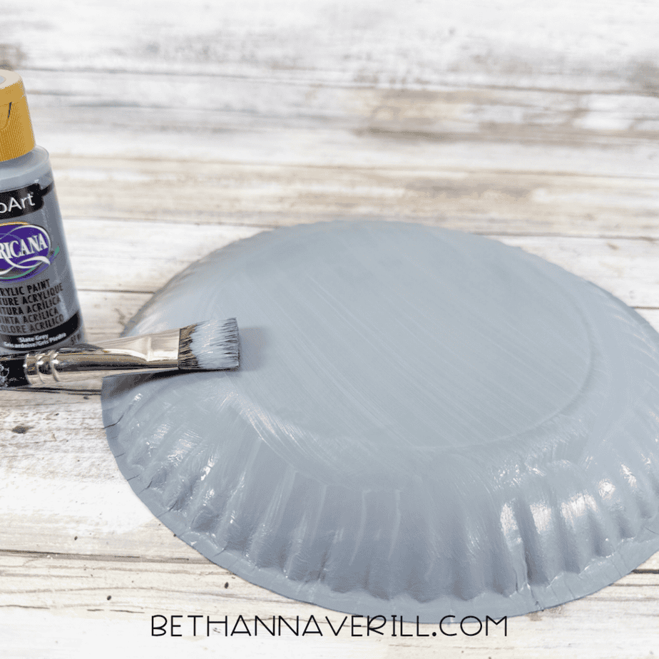 Paintbrush applying gray acrylic paint to the back of a paper plate, with a gray paint bottle beside it.