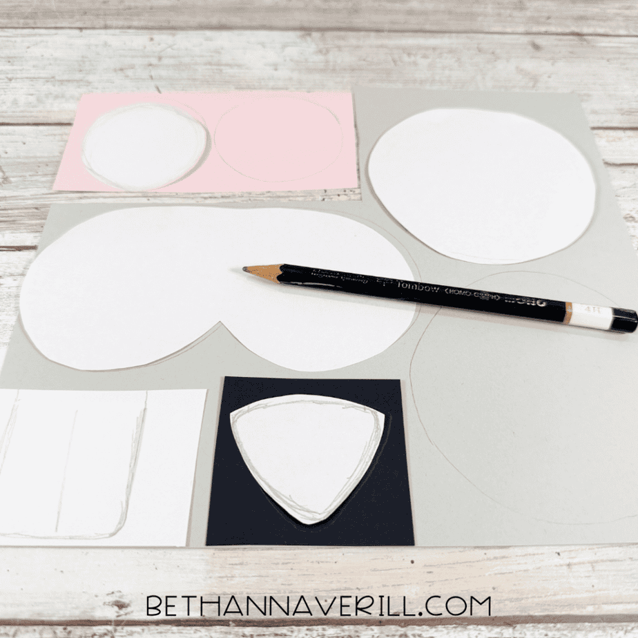 White paper templates arranged on gray, pink, black, and white paper with traced outlines and a black pencil.