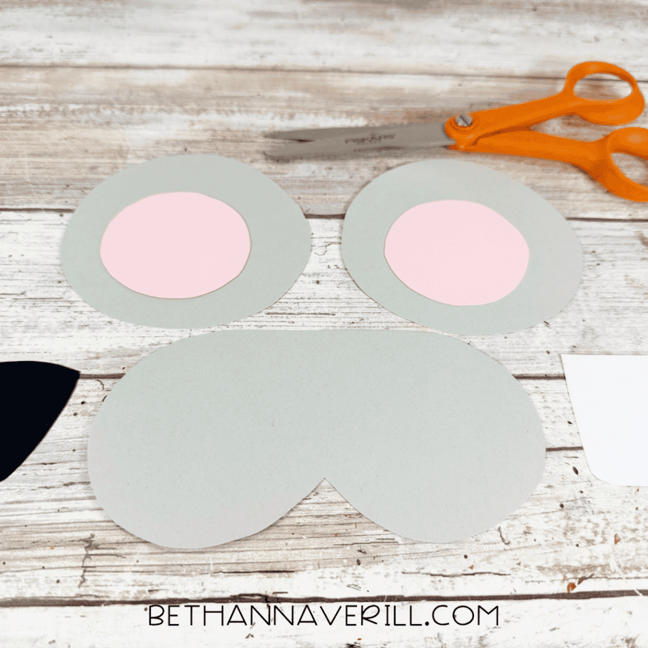 Cut gray and pink circular paper pieces, a large gray muzzle shape, black paper, and a white rectangular piece arranged beside orange scissors.
