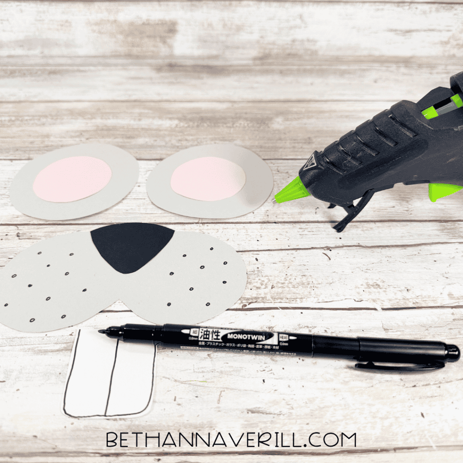 Gray muzzle piece with a black triangular nose and small black dots, two gray ears with pink centers, and a white tooth piece beside a hot glue gun and black marker.